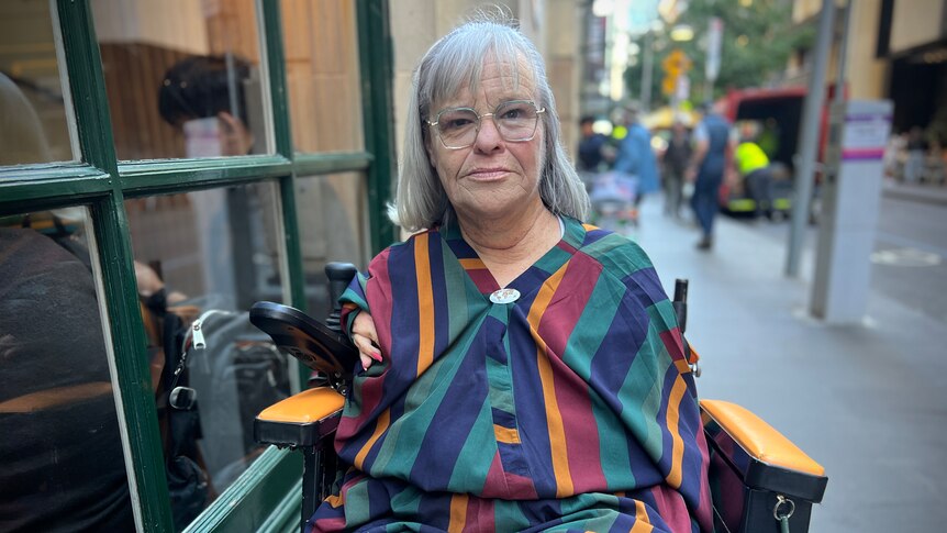A middle aged white woman without a right arm sits outside in a motorised wheelchair, wearing a striped shirt and big glasses. 