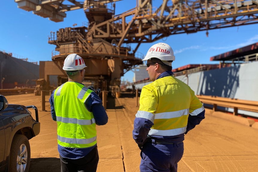 Rio Tinto workers stand talking on the dock at Dampier with loader in front of them and ships to the side.