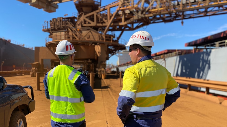 Rio Tinto workers stand talking on the dock at Dampier with loader in front of them and ships to the side.