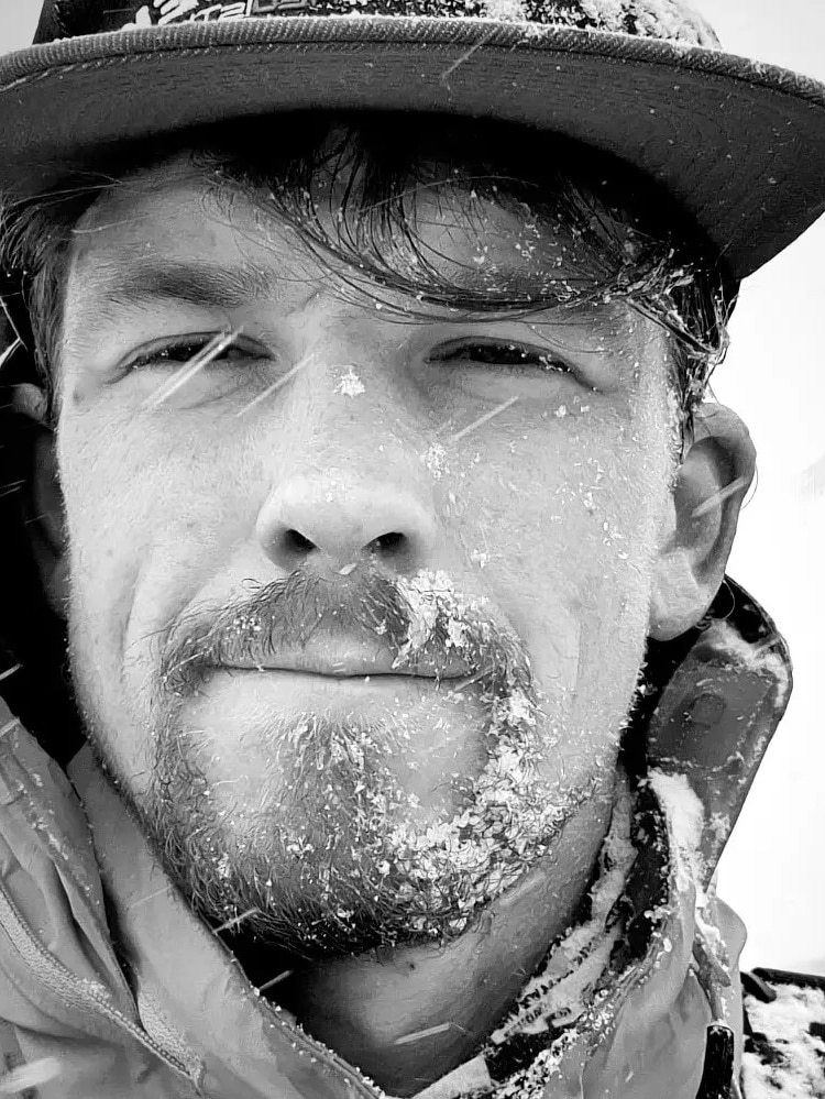 Close shot of a man's bearded face as he squints through falling snow.