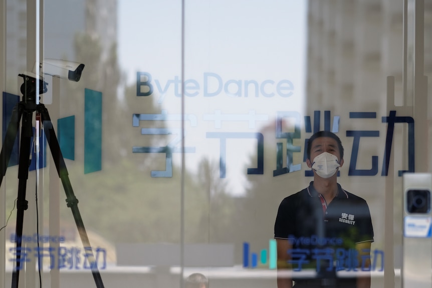 A Chinese man wearing a polo shirt and a facemask stands behind a glass office door beside a security ca