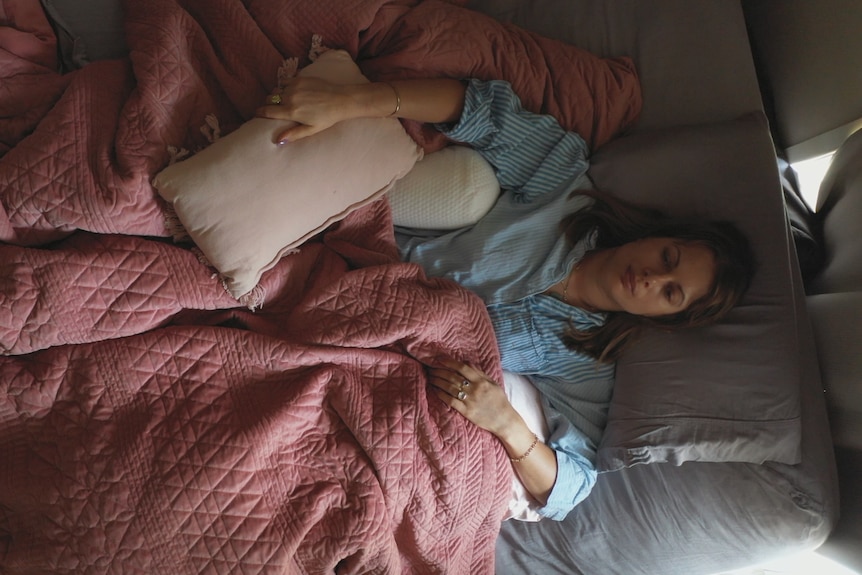A woman lying in a bed with a pink bedspread holding a pillow