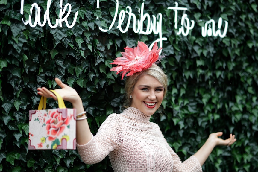 A woman, wearing a fascinate in front of a garden wall, at a horse racing meet, holding a handbag