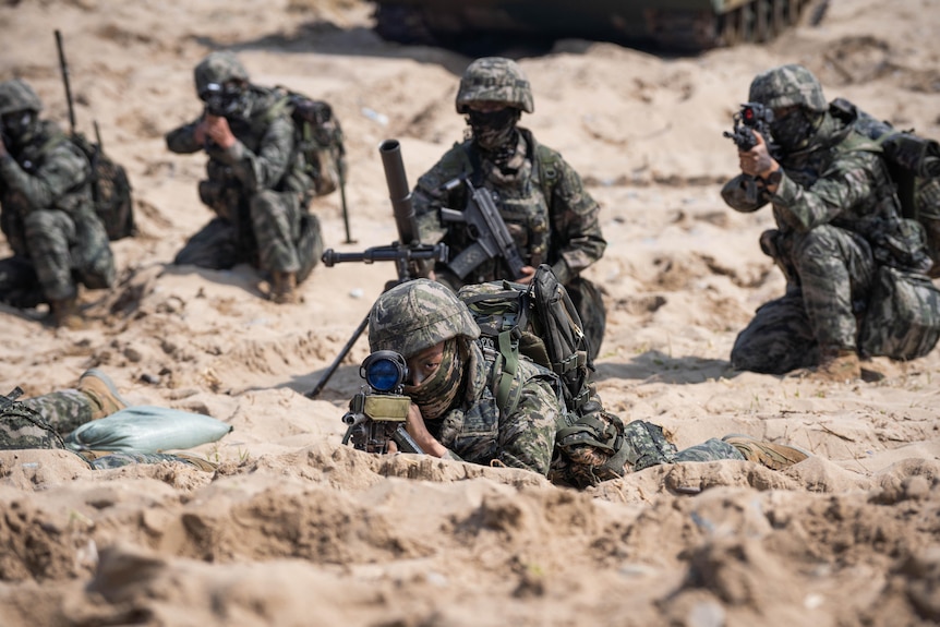 Marines in camouflage gear crouch on a beach, aiming weapons towards camera