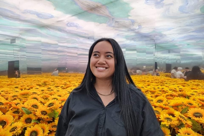 Woman with long dark hair wearing black buttoned shirt stands in room of yellow sunflowers. 