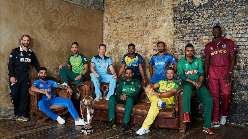 Ten cricket players wearing different coloured clothing stand or sit on brown leather sofas against a bare-brick background