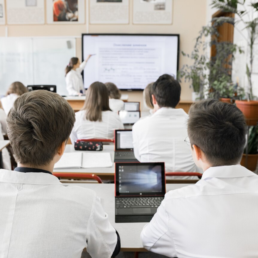 Rows of students wearing collared uniforms and using small laptops at their desks, while a teacher points to a whiteboard.