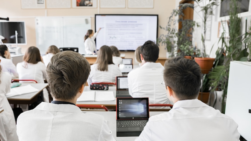 Rows of students wearing collared uniforms and using small laptops at their desks, while a teacher points to a whiteboard.