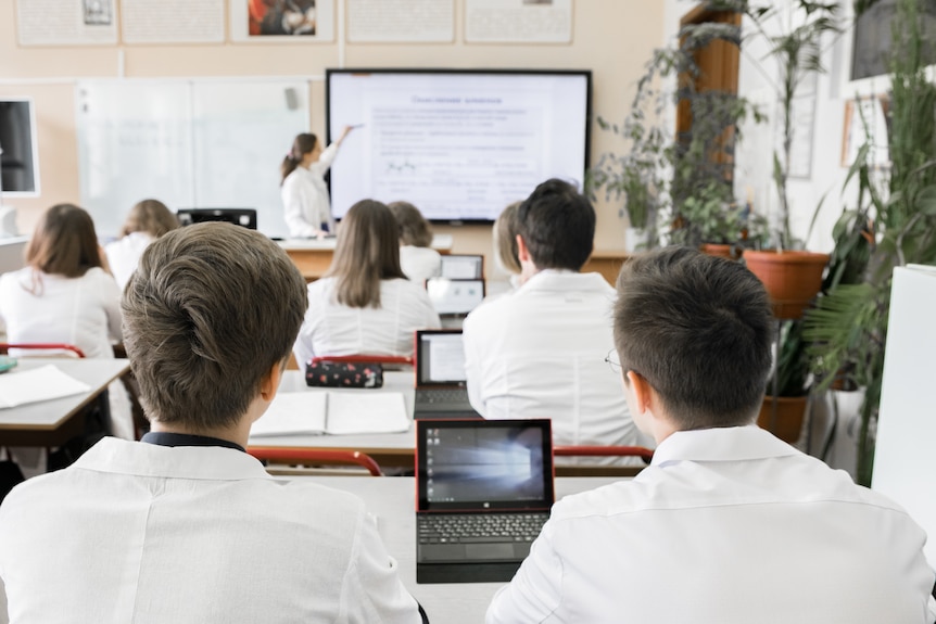 Rows of students wearing collared uniforms and using small laptops at their desks, while a teacher points to a whiteboard.
