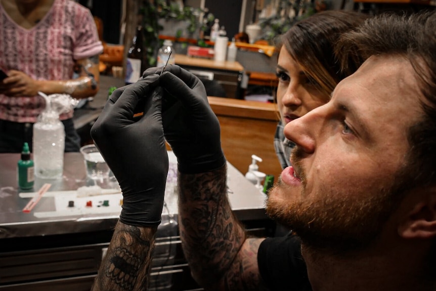 A man holds up a tattoo needle in his hands, whilst wearing gloves. A women looks on next to him. 