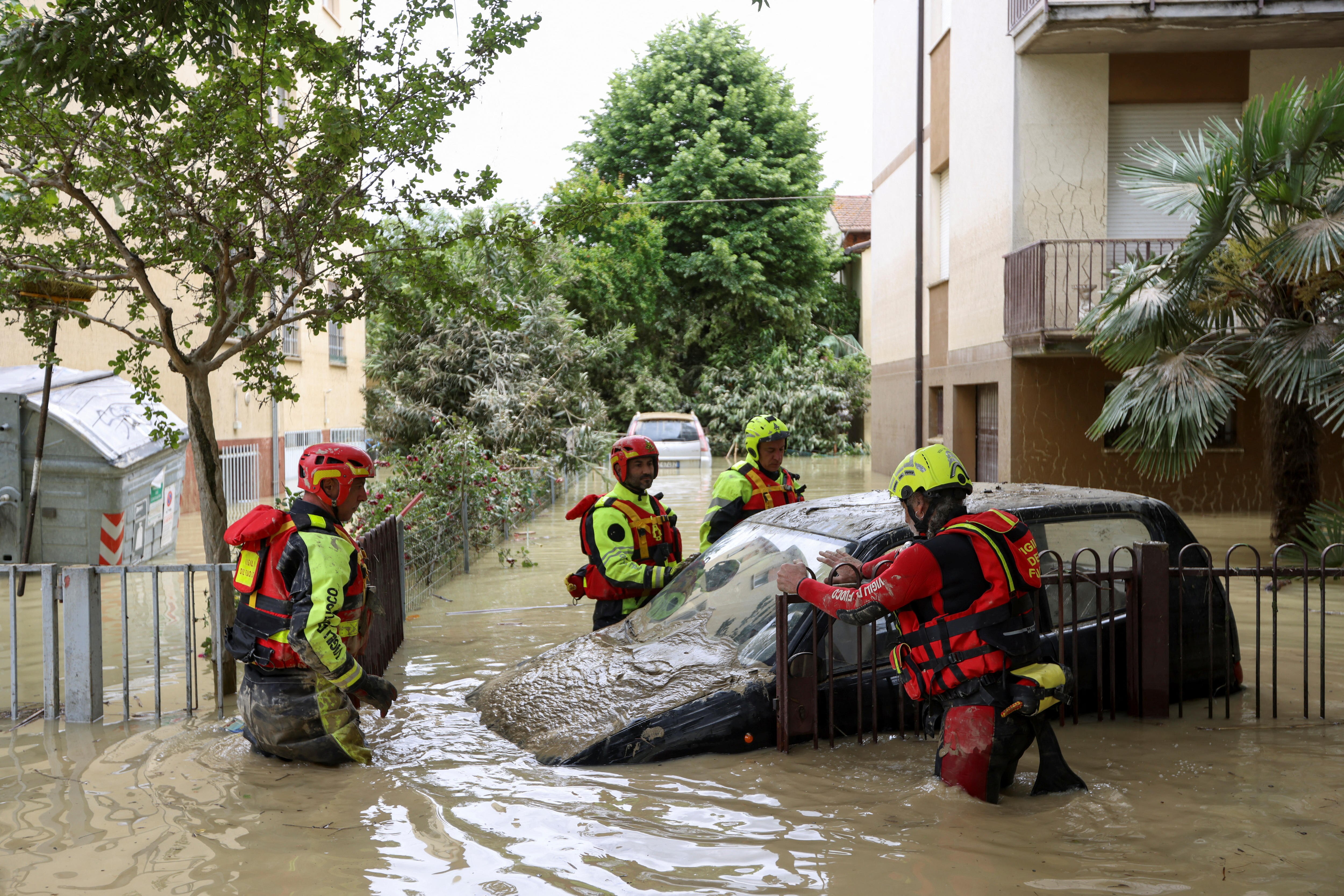 Rescuers in red and yellow outfits stand near a car partially submerged in muddy waters. 