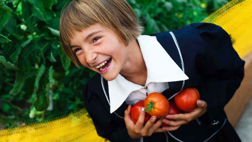A young child smiles in a garden holding homegrown tomatoes she has harvested, easy veggies to grow with kids.