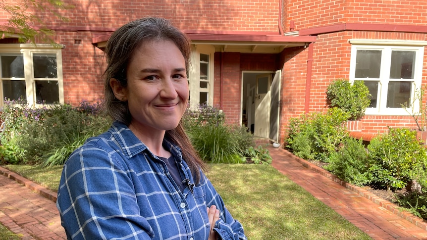 A woman in a blue flannel stands in front of a brick home.