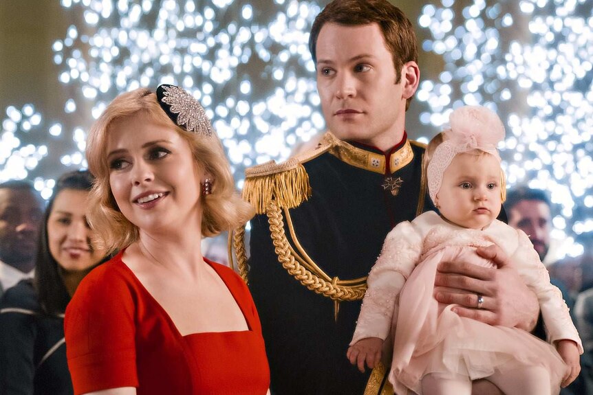Rose McIver in a red dress and Ben Lamb in royal outfit, holding a baby with a headband, in The Princess Switch: Switched Again.
