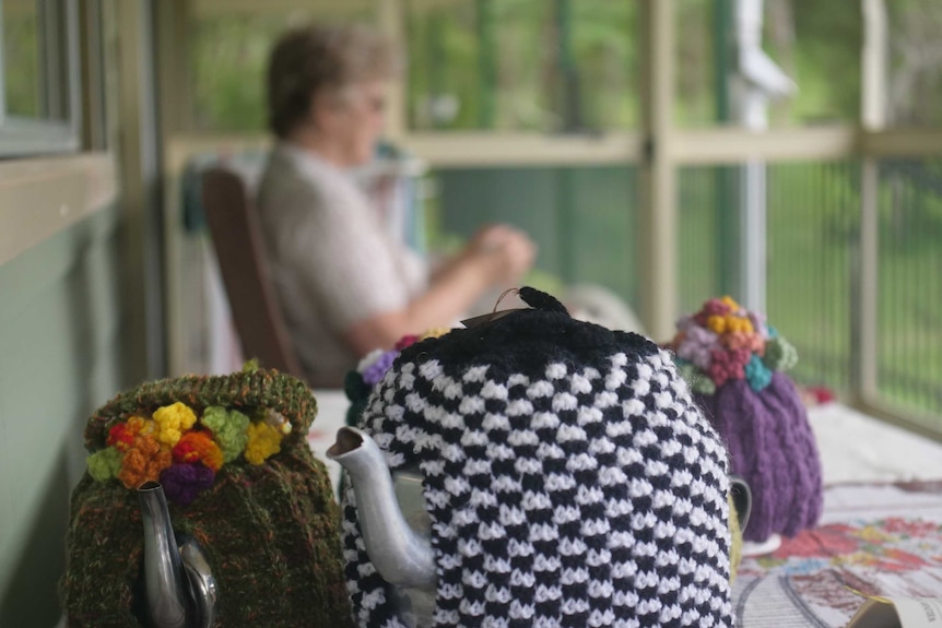 A black and white knitted tea cosy on a tea pot, with a woman knitting in the background.