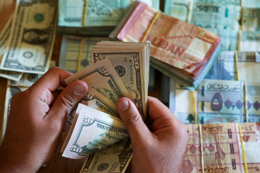 A man's hands peel through US dollar bank notes on a table, with stacks of international currencies below