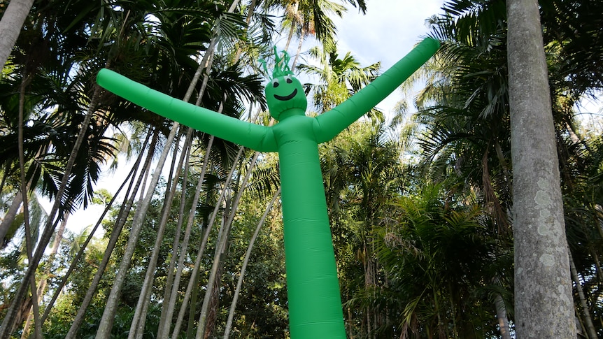 A bright green inflatable wavy man stands among a tree canopy