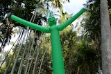 A bright green inflatable wavy man stands among a tree canopy