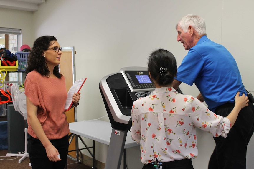 Woman talks to patient on running machine, as physio places hand on patient to support him