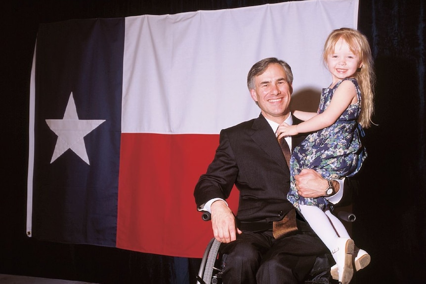 Greg Abbott sits in his wheelchair with his daughter on his lap in front of a big Texas flag 
