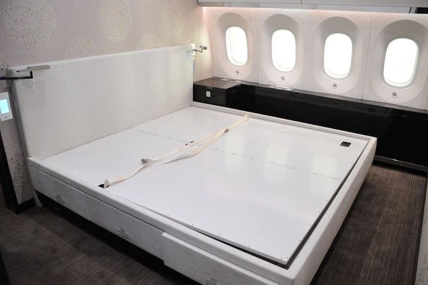 A white bed with a headboard inside a plane, with plane windows running next to it