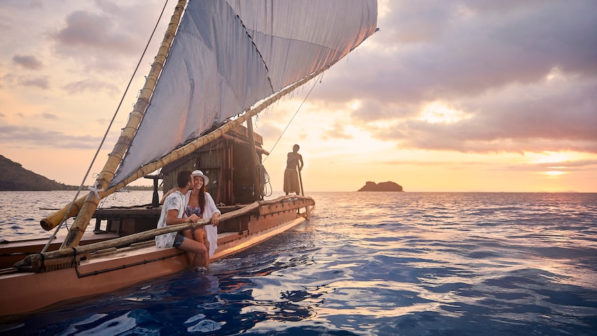 A man and a woman sit on a Fijian sailboat with a sunset in the background.
