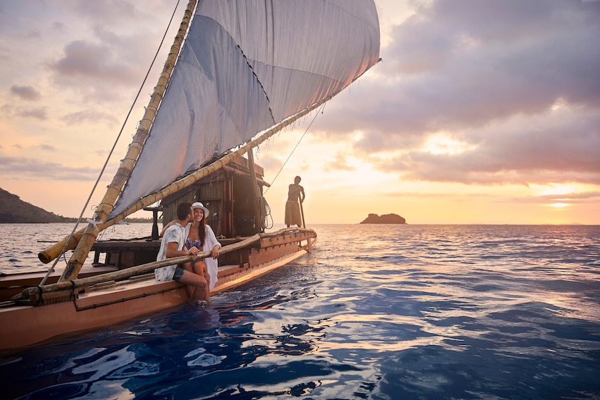 A man and a woman sit on a Fijian sailboat with a sunset in the background.