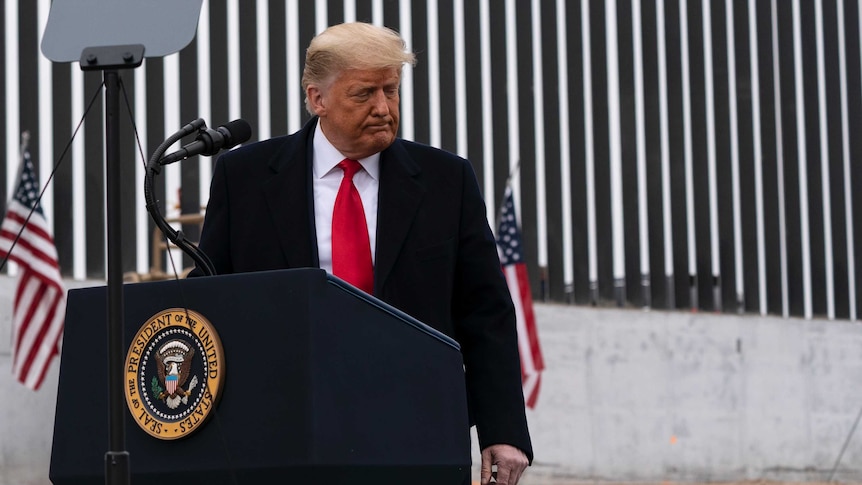 Donald Trump stands at a lectern in front of a corrugated wall.