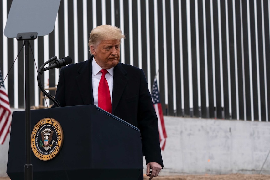 Donald Trump stands at a lectern in front of a corrugated wall.