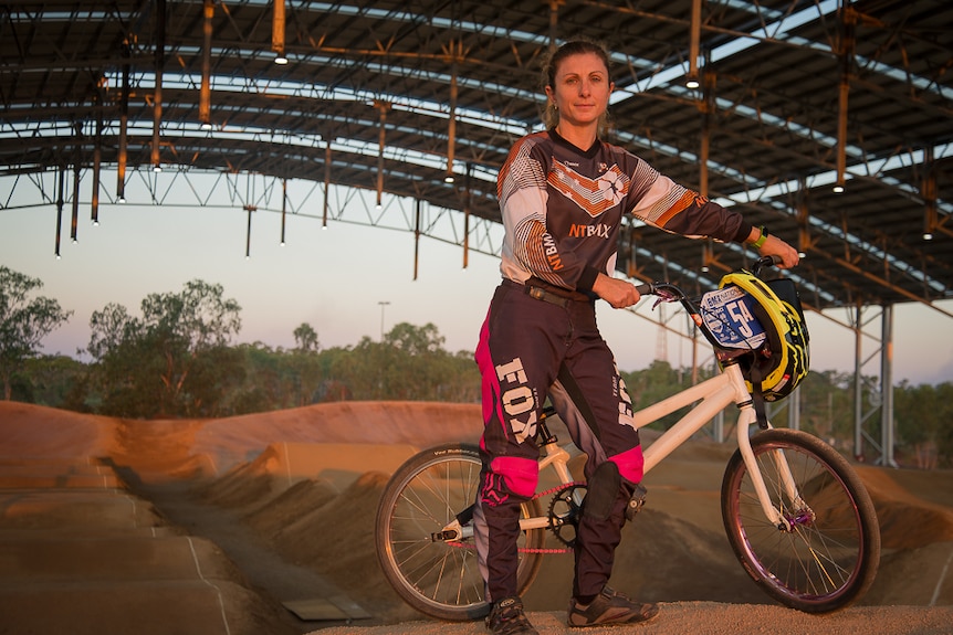 A middle aged woman stands at the highest point of a BMX track with her white bike and fluorescent yellow helmet.