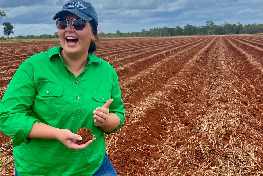 A young Indigenous woman with a green button-up shirt, hat and sunglasses poses on a farm smiling.
