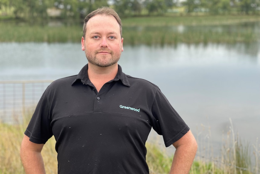 A man in a black t-shirt stands in front of a farm dam 