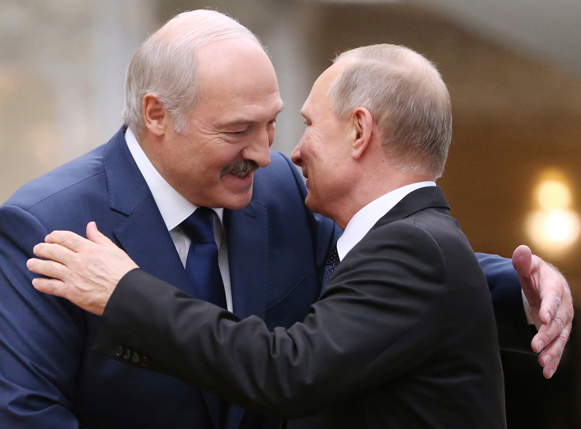 Alexander Lukashenko in blue suit smiles while leaning down to hug Vladimir Putin in black suit