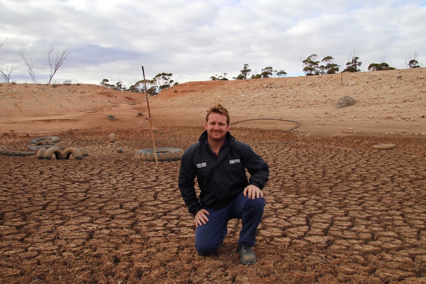 A man kneels in a dry dam on cracked ground with a pole behind him showing the normal water level.