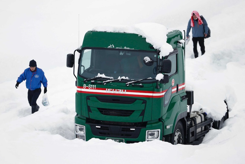 Two people wade through the deep snow past a green truck stuck in the snow.