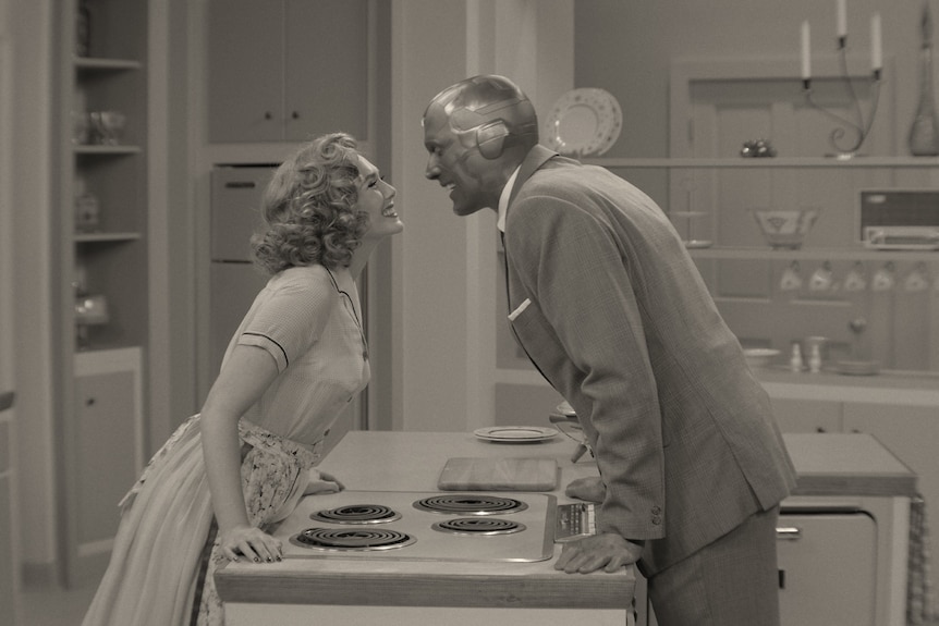 A 30-something woman and a masked man in 50s-style clothes smile lovingly at each other while leaning on the kitchen counter