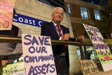 A man in a dark suit looks out at crowd, surrounded by signs some read save our community assets