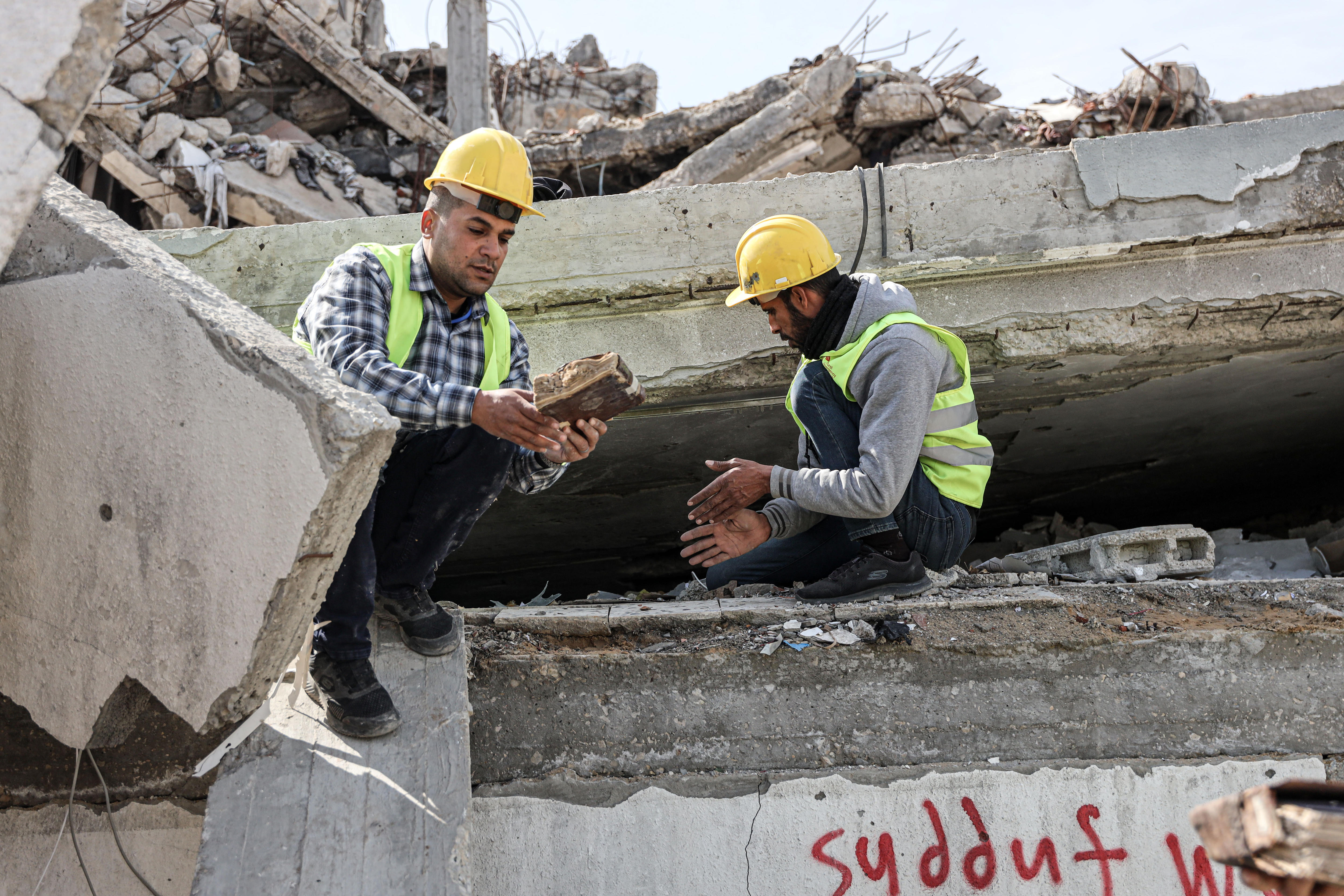 Two men wearing hard hats and yellow construction jackets hold items retrieved from the rubble of a building.