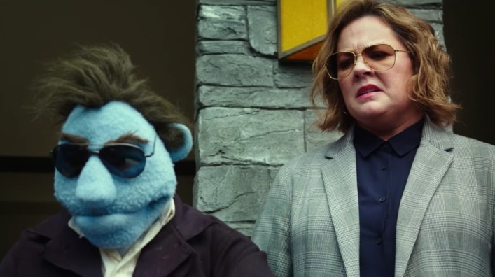 Melissa McCarthy, with aviator sunglasses, stands behind a blue puppet in similar clothing. She is grimacing