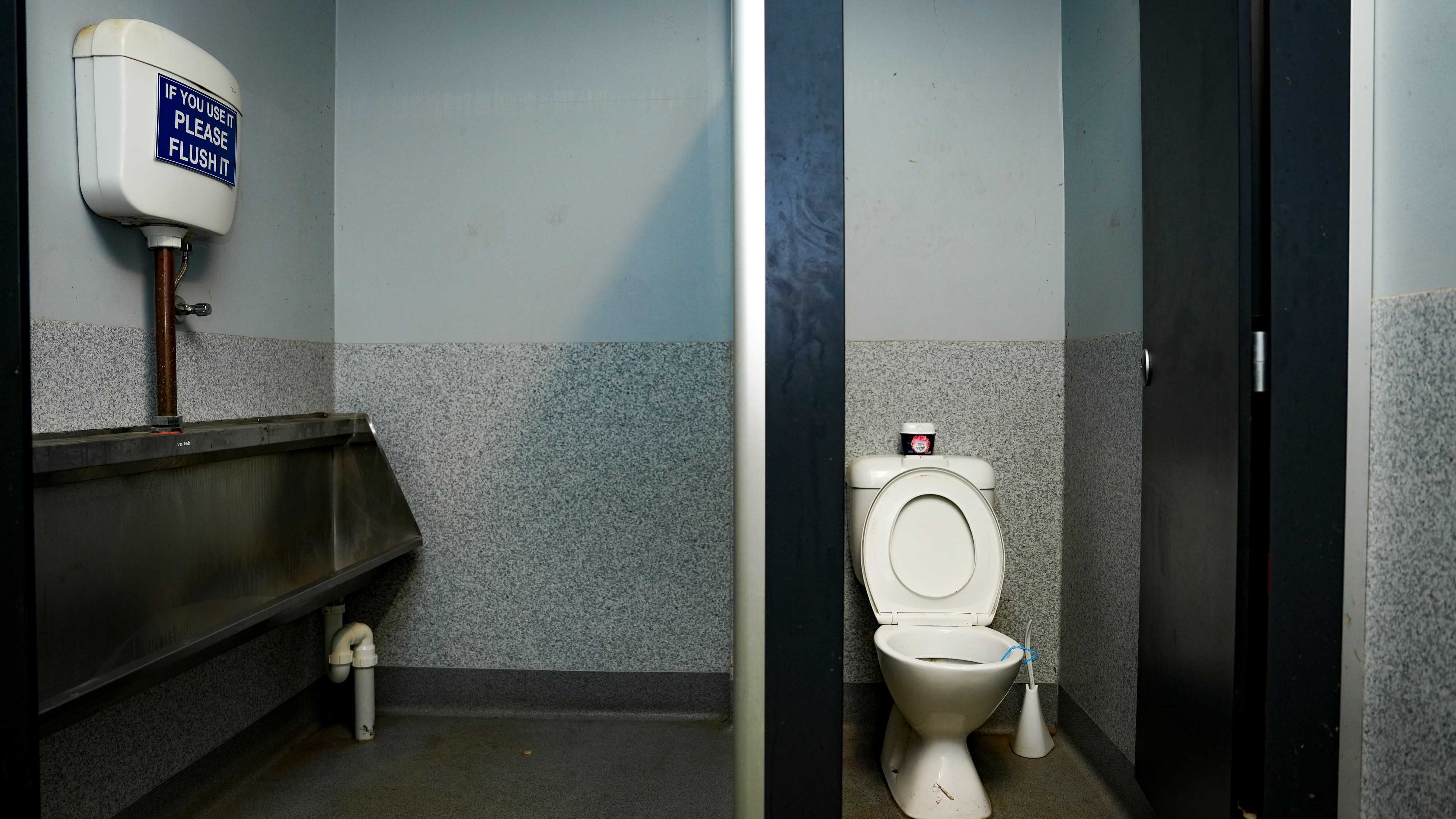 A toilet cubicle next to a wall with a urinal along it.