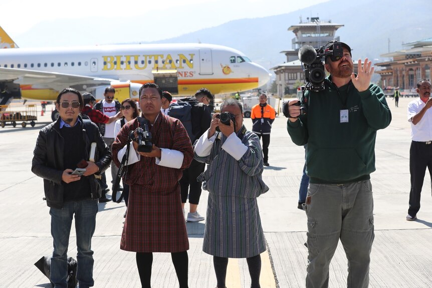 Smith filming with camera and waving, standing next to local media in traditional Bhutan dress on airport tarmac.