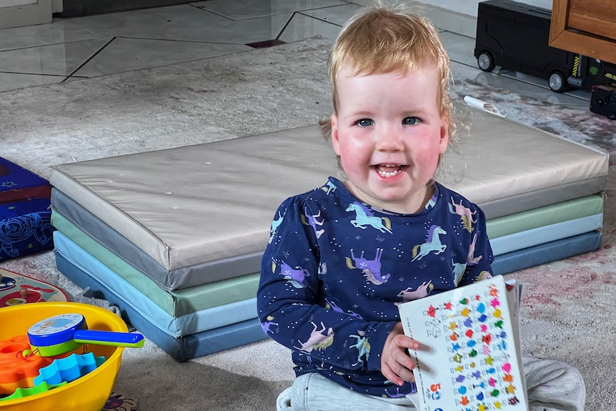 A baby sitting up, smiling, with a book in her hands.