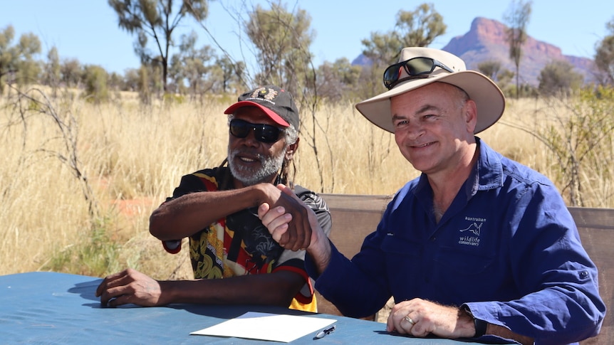 Two men sit at a table in the desert, shaking hands