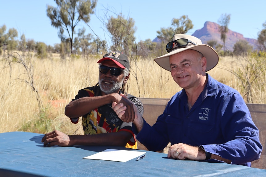 An indigenous man and white man sit at a table in grassland, shaking hands