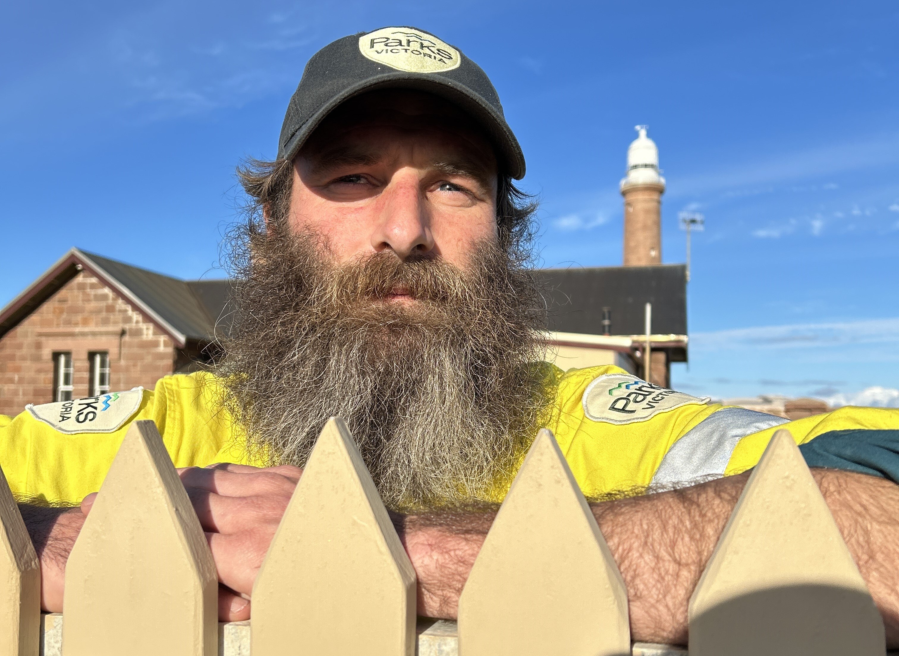 A man with a long beard and wearing a cap, leans on a picket fence with a stone house and lighthouse behind him.