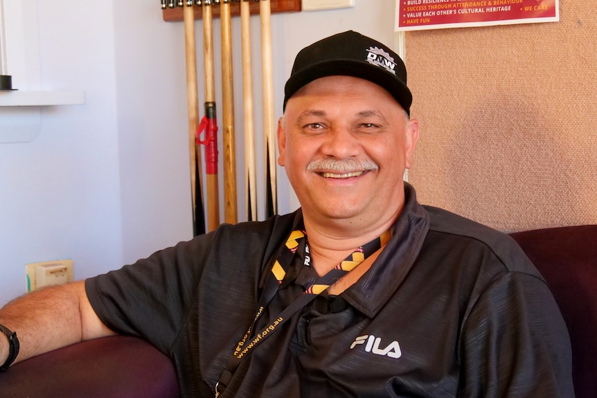 A seated, smiling man, facing the camera. He is wearing a black, short-sleeved shirt and black cap.