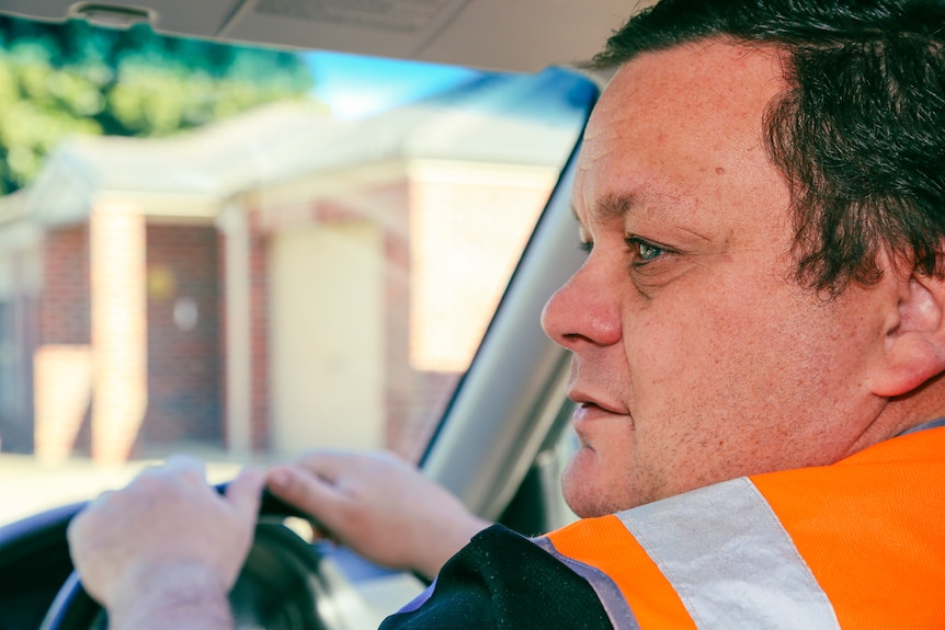 A man sits behind the wheel of his car, seen from behind, and looks to the left of frame.