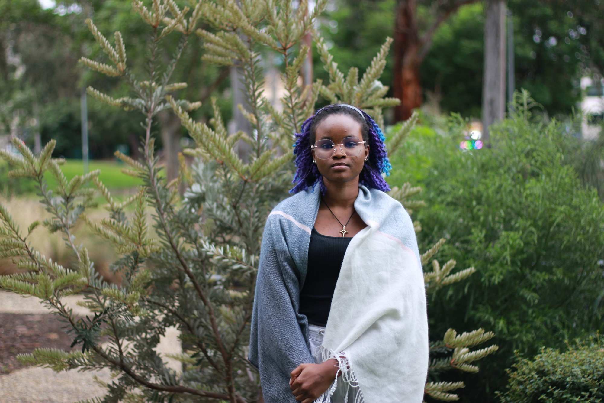 A young woman wears a shawl over her shoulders standing in a garden on a grey day.
