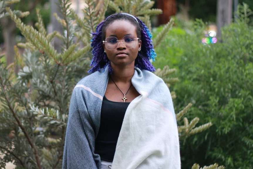 A young woman wears a shawl over her shoulders standing in a garden on a grey day.