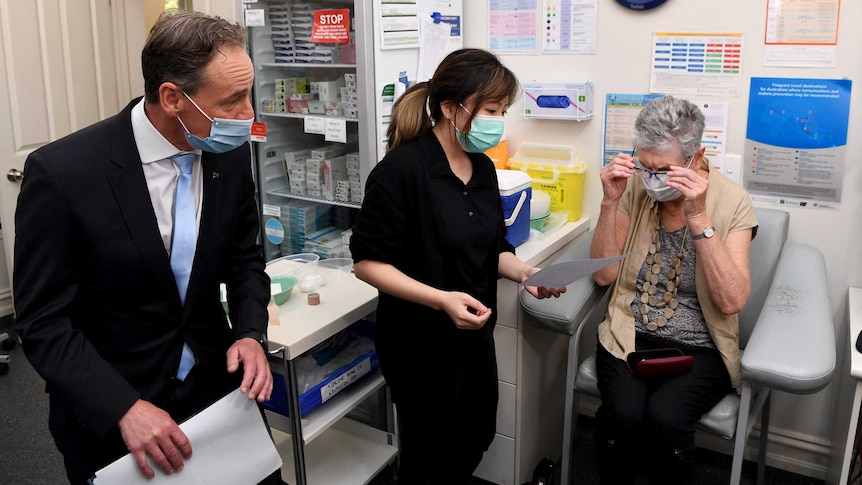 man on left in suit and mask - Greg Hunt - plus nurse and elderly woman getting injection